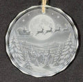 A round glass Christmas ornament featuring an etched winter night scene with Santa Claus in his sleigh, reindeer, a full moon, snowy mountains, and pine trees. The ornament hangs from a tan string against a dark textured background.